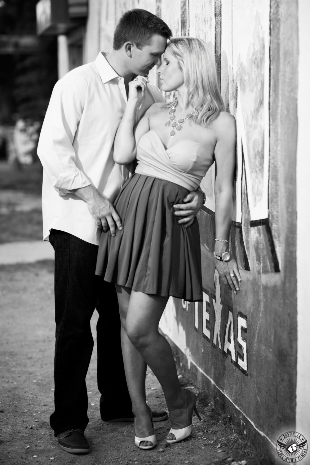 Sexy blond girl wearing an a-line off white and green dress and white high heels and beaded necklace nuzzles and gently touches the chin of a guy with dark hair wearing a white untucked button up dress shirt and dark jeans in front of the Greetings From Austin sign near SOCO in this impactful engagement portrait in South Austin.  